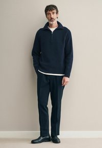 Pulover bleumarin cu nervuri, cu guler înalt, asortat cu pantaloni închiși la culoare și pantofi negri. Materialul pare moale, texturat și ușor ajustat.