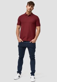 Tummanpunainen polo-paita lyhyillä hihoilla, kolmella napilla varustettu kaulus ja hienovarainen logo; yhdistetty tummansinisiin slim fit -farkkuihin ja valkoisiin lenkkitossuihin.