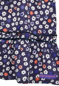 Dunkellila Stoffkleid mit weißen Blumen und Smiley-Gesichtern. Gerüschter Saum mit schwarzem Paillettenbesatz und einem "KEEP ON SMILING"-Label.
