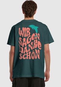 Mørk teal t-shirt med oversized, legende tekst i pink: "WIR SAGEN DANKE SCHÖN" med en teal delfin accent. Fremstillet af blødt stof, afslappet pasform.
