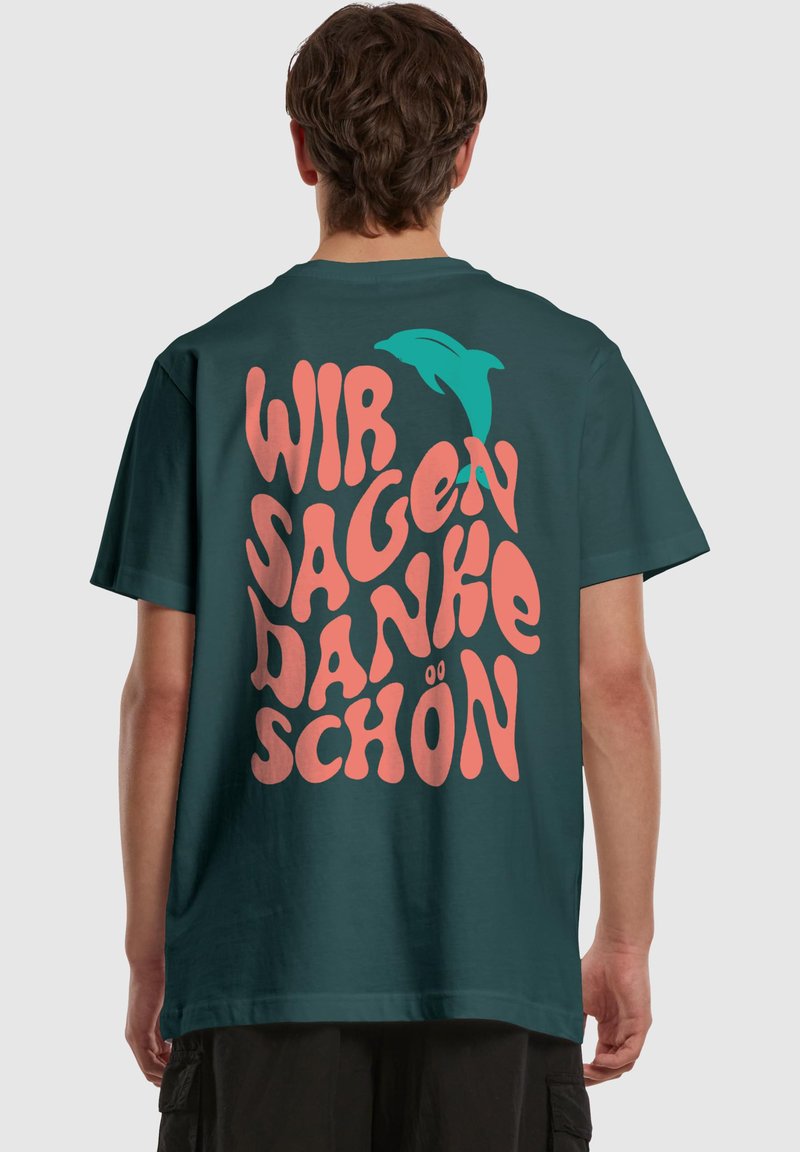 Mørk teal t-shirt med oversized, legende tekst i pink: "WIR SAGEN DANKE SCHÖN" med en teal delfin accent. Fremstillet af blødt stof, afslappet pasform.