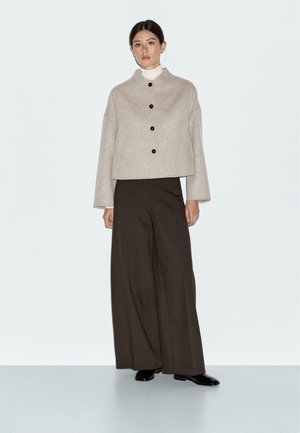 Beige cropped jacket with a high collar and black buttons, paired with loose, dark brown wide-leg pants. Smooth fabric, minimalistic design.