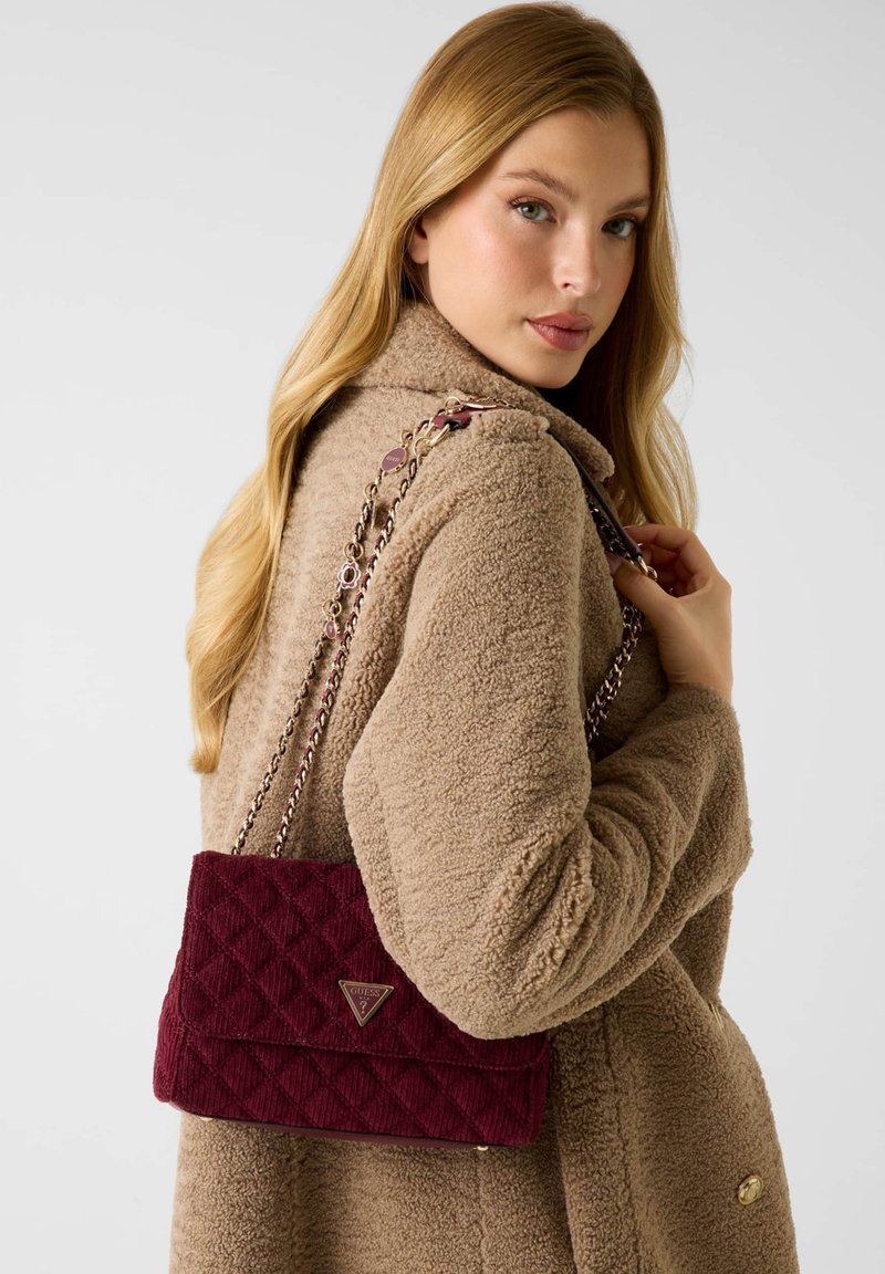 Młoda kobieta z długimi blond włosami, nosząca beżowy płaszcz teddy, trzymająca pikowaną burgundową torbę na ramię z złotymi łańcuszkami.