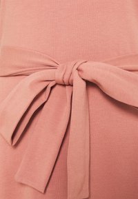 Robe en coton d'un rose doux avec une ceinture nouée à la taille. Le tissu présente une texture lisse et un léger stretch, mettant en valeur la silhouette.