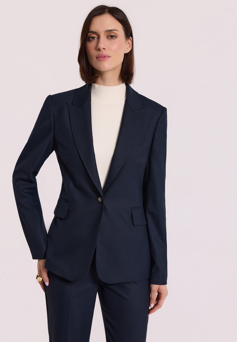 Navy blazer z dopasowanymi ramionami, zapinany na jeden guzik, z dwoma przednimi kieszeniami. Noszony na kremowym golfie, o gładkiej fakturze.