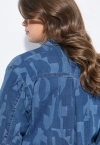 Giacca di jeans di colore blu medio con un sottile motivo astratto, colletto classico e pattina con bottoni, caratterizzata da una silhouette rilassata.