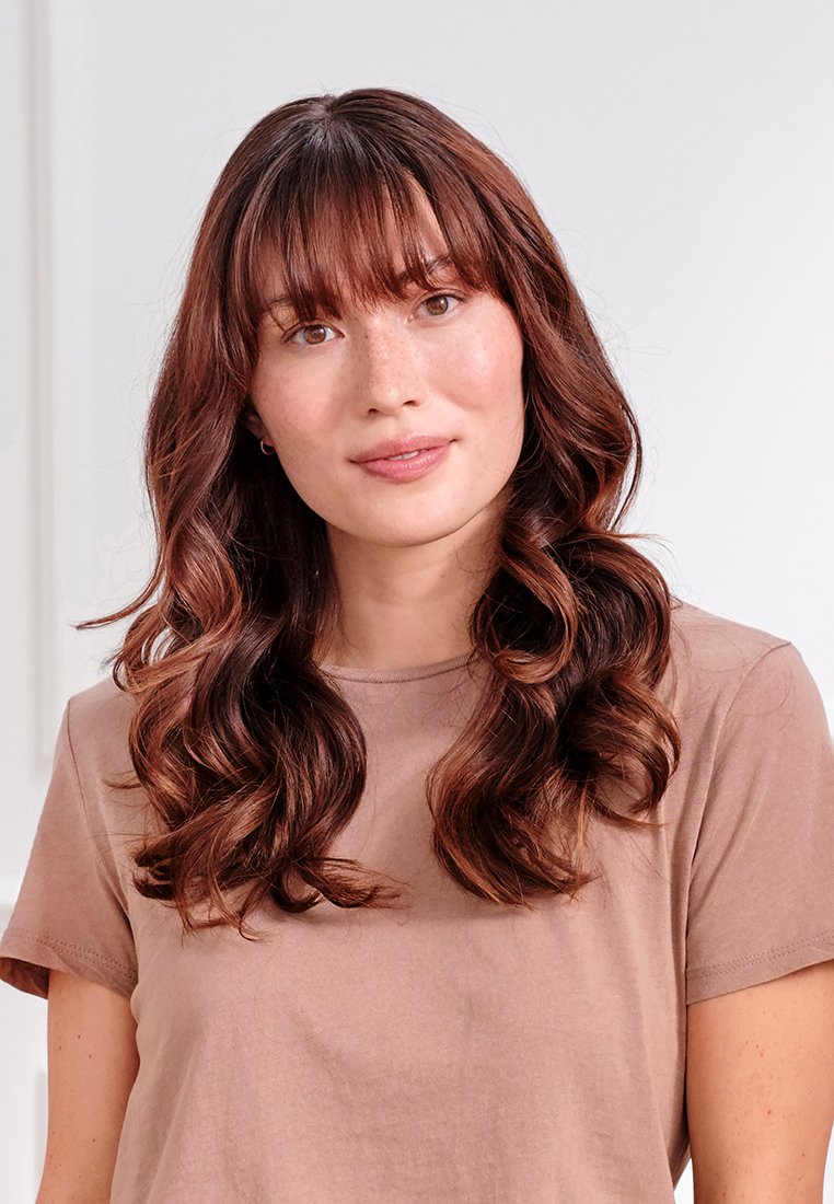 Długie, falujące ciemne brązowe włosy z grzywką, noszące jasnobrązowy T-shirt. Gładka skóra z piegami, neutralne tło.