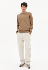 Brauner Strickpullover, kombiniert mit hellen, locker sitzenden Hosen und grauen Sneakers. Das Model steht mit den Händen in den Taschen vor einem neutralen Hintergrund.