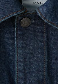 Cuello de chaqueta de mezclilla azul oscuro con costuras naranjas y un botón metálico negro con la inscripción "MNG DENIM".
