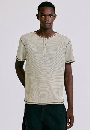Jeune homme portant un t-shirt henley à manches courtes, rayé de couleur claire, et un pantalon foncé, debout devant un fond uni.