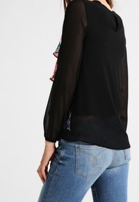 Blouse noire avec manches longues transparentes et accents de motif floral sur les poignets, dotée d'une ouverture en forme de goutte dans le dos et d'une coupe décontractée. Associée à un jean bleu.