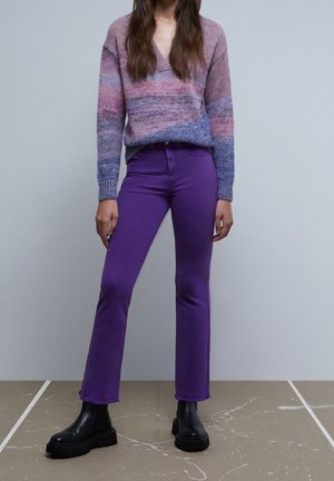 Femme portant un pull en laine dégradé violet, un pantalon violet droit et des bottines Chelsea noires épaisses, debout sur un sol texturé.
