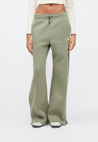 Άτομο που φοράει χακί φαρδιά παντελονιού φόρμας με κορδόνι στη μέση και σκισίματα στα τελειώματα, συνδυασμένο με λευκά αθλητικά παπούτσια και λευκό κοντό μπλουζάκι με μακριά μανίκια.