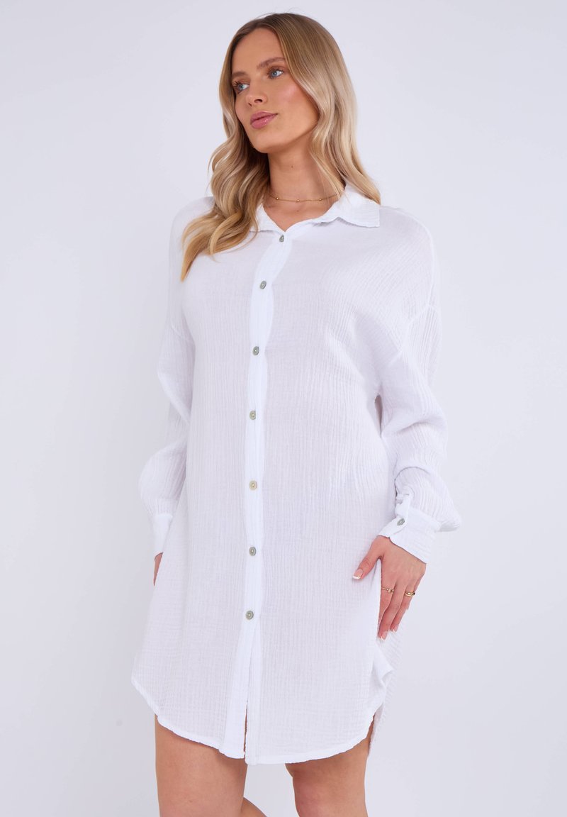 Robe blanche à boutons, en tissu texturé avec des manches longues, mettant en avant un col de chemise, des boutons dorés et des fentes latérales à l'ourlet.