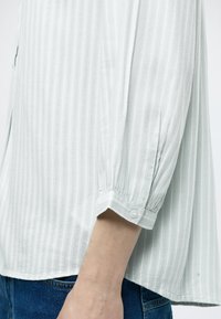 Chemise gris clair à manches longues avec des rayures verticales et des poignets boutonnés portée avec un jean bleu sur le bras et la taille d'une personne.