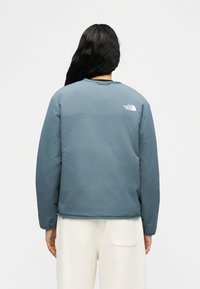 Άτομο με μακριά σκουρόχρωμα μαλλιά που φοράει μπλε μπουφάν The North Face και άσπρο παντελόνι, εμφανιζόμενο από πίσω με φόντο απλό.