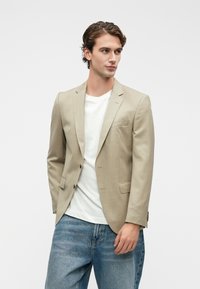 Μπεζ σακάκι με κομψή εφαρμογή, μονής σειράς, με κανονικά πέτα και δύο μπροστινές τσέπες, φορεμένο πάνω από λευκή μπλούζα και μπλε τζην.