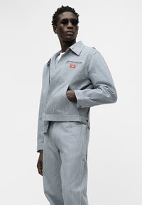 Άνδρας ντυμένος με μπλε και λευκή ριγέ ζακέτα και παντελόνι που ταιριάζει, φορά γυαλιά ηλίου, κοιτάζοντας πλάγια με το χέρι στην τσέπη της ζακέτας.