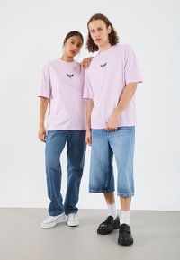 Light pink oversized t-shirts with black butterfly graphic. Model wears blue jeans and white sneakers, while the other has denim shorts and black shoes.