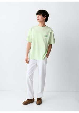 Jonge man in een lichtgroen T-shirt met print, witte broek en bruine schoenen, staand tegen een witte muur, kijkt opzij met handen in de zakken.