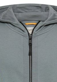 Grijze hoodie met rits, voorzien van een capuchon, gladde textuur, centrale ritssluiting met zwarte hardware, en een label met oranje accent aan de binnenkant van de kraag.
