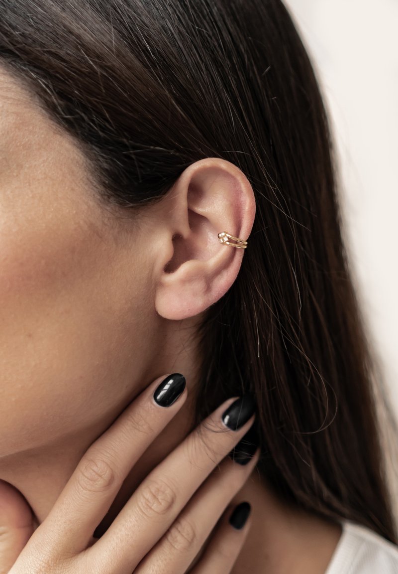 Goldener Ohrcuff mit drei Bändern und einem Kristallakzent, getragen am oberen Ohr. Die glatte Metalloberfläche steht im Kontrast zu den schwarzen Nägeln.