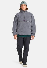 Grijze fleece trui met een kwartrits, voorzien van een patroonband over de borst en zijzakken. Gecombineerd met een donkere broek en sneakers.