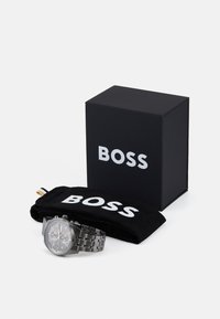 Melns hronogrāfa pulkstenis ar nerūsējošā tērauda siksniņu, novietots uz melna maisiņa un kastes, abiem ar baltu "BOSS" zīmolu.