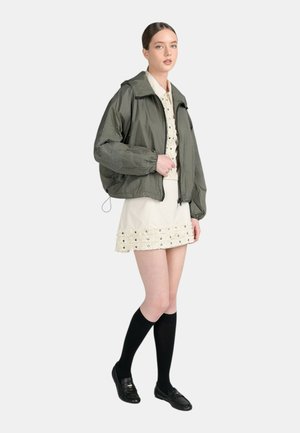 Donna che indossa una giacca bomber verde sopra un abito crema ricamato, calzini neri al ginocchio e mocassini neri, in piedi davanti a uno sfondo uniforme.