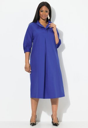 Abito blu, alla lunghezza del ginocchio, con colletto, maniche a tre quarti e pieghe frontali. Indossato con scarpe nere con dettagli decorativi.