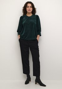 Blusa in velluto a coste verde scuro con maniche a tre quarti, abbinata a pantaloni neri attillati e stivaletti neri. Design minimalista.