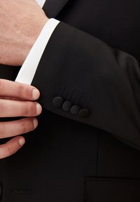 Svart kavajärm med en vit manschett, prydd med fyra matta svarta knappar och dekorativ söm nära manschetten.