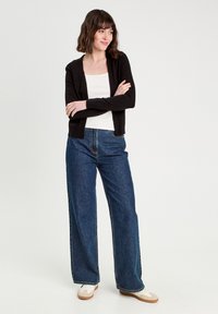 Cardigan noir par-dessus un débardeur blanc, associé à un jean bleu ample. La tenue présente une coupe décontractée et une texture côtelée subtile.