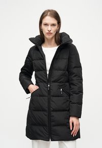 Γυναίκα φορώντας μακρύ μαύρο καπιτονέ μπουφάν με ψηλό γιακά, λευκό τοπ και λευκό παντελόνι, στέκεται μπροστά σε απλό λευκό φόντο.