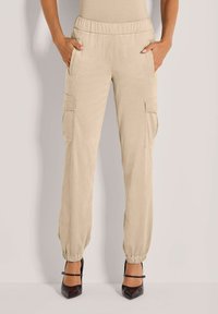 Beige Cargo-Hosen mit elastischem Bund und Bündchen, seitlichen Klappentaschen, getragen mit schwarzen spitzen High Heels, Hände in den vorderen Taschen.