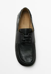 Chaussure en cuir noir avec une texture lisse, bout rond et laçage décoratif. L'intérieur présente une doublure beige avec l'impression de la marque.