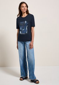 T-shirt marine avec un design graphique d'un visage et de feuillage, associé à un jean large bleu clair et des sandales foncées sur un fond neutre.