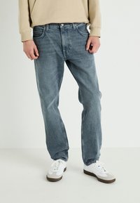 Jeans de denim azul claro con corte recto, con bolsillos estándar y un ligero desgaste. Combinados con zapatillas blancas y una blusa beige.