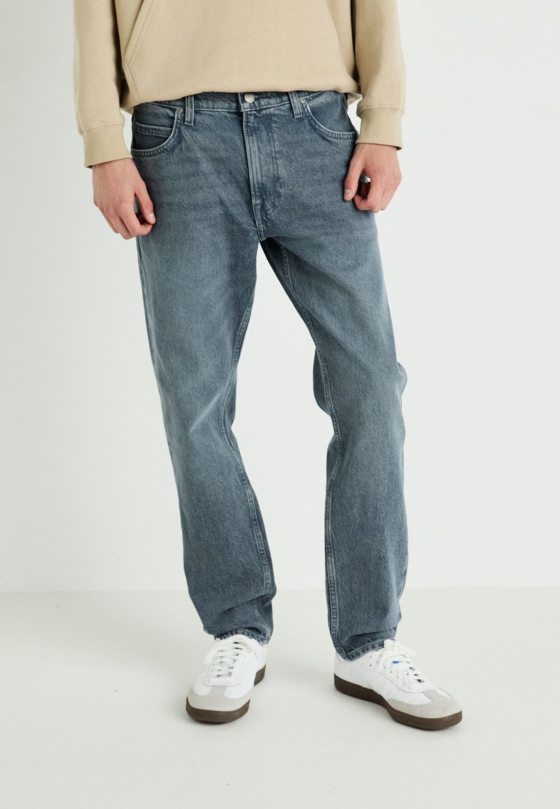 Jeans de denim azul claro con corte recto, con bolsillos estándar y un ligero desgaste. Combinados con zapatillas blancas y una blusa beige.
