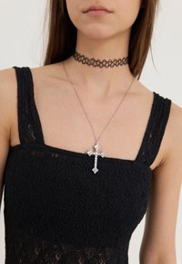 Fekete csipkés top vastag pántokkal, ezüst színű nyaklánccal, amelyen egy nagy díszes kereszt medál található, gyöngyökkel díszítve.