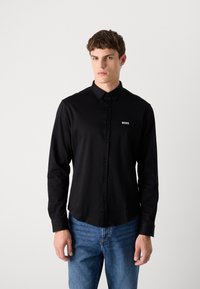 Black long-sleeve shirt with button-down collar, featuring a small white "BOSS" logo on the left chest, paired with light blue jeans.