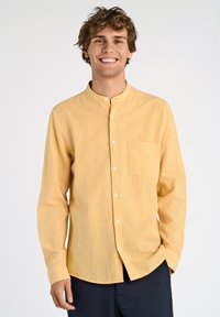 Camicia gialla a maniche lunghe realizzata in tessuto texturizzato, con colletto rialzato, tasca frontale e bottoni bianchi. Indossata con pantaloni scuri.