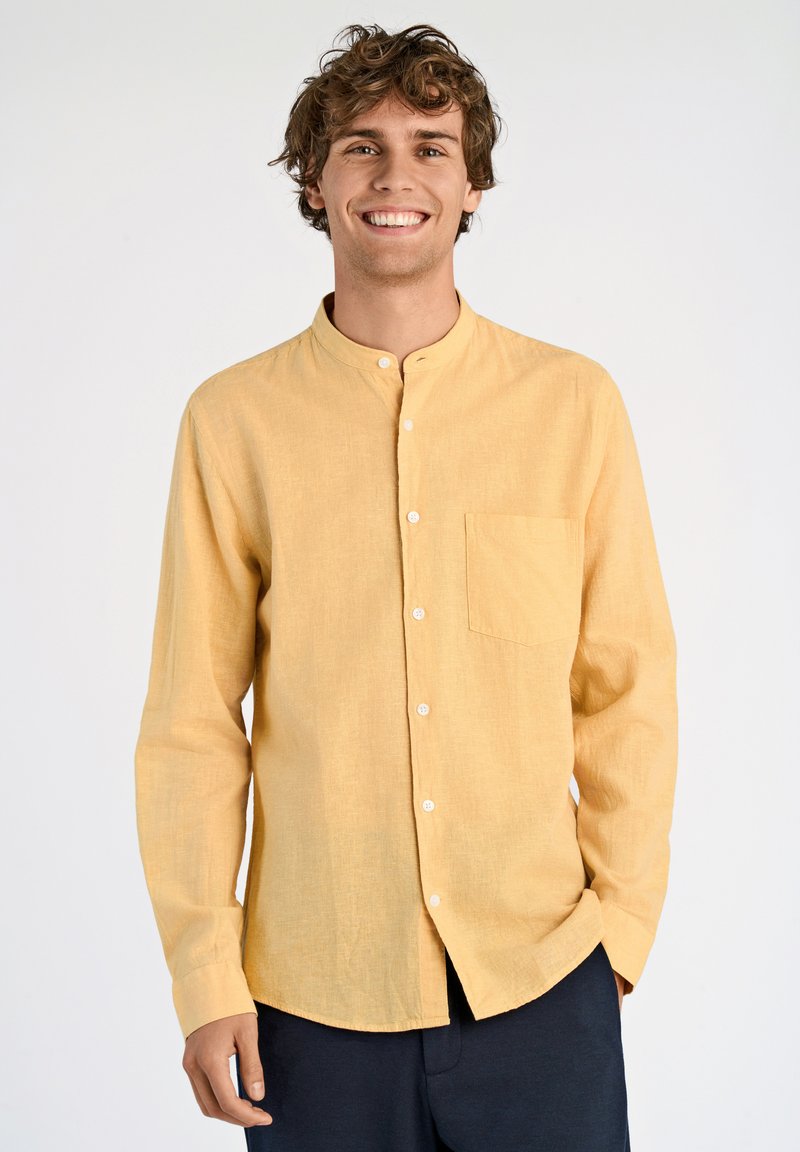 Camicia gialla a maniche lunghe realizzata in tessuto texturizzato, con colletto rialzato, tasca frontale e bottoni bianchi. Indossata con pantaloni scuri.