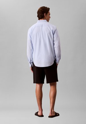 Mężczyzna zwrócony tyłem, ubrany w jasnoniebieską koszulę z długim rękawem, czarne szorty i czarne klapki na jednolitym szarym tle.