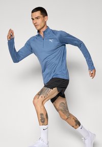 Tricou atletic de culoare albastru deschis, cu mânecă lungă și fermoar pe un sfert, combinat cu pantaloni scurți negri. Tatuaje notabile pe picioare. Șosete albe și adidași vizibili.