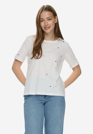 Młoda kobieta o długich włosach, ubrana w białą koszulkę z małymi kolorowymi kropkami oraz niebieskie jeansy, stoi z rękami za plecami.