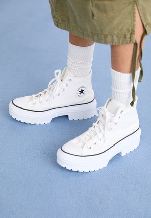Białe wysokie masywne trampki Converse noszone z białymi prążkowymi skarpetami i oliwkową spódnicą stojące na jasnoniebieskiej podłodze.