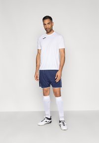 Balti sportiniai marškinėliai su juodu logotipu, derinami su tamsiai mėlynais šortais su baltais dryžiais, baltais iki kelių siekiančiais kojiniais ir juodais bei baltais futbolo bateliais.