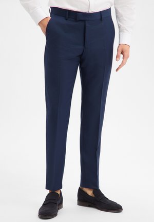Mann trägt marineblaue, maßgeschneiderte Hose, weißes Hemd und schwarze Wildleder-Loafer, steht mit einer Hand in der Hosentasche vor weißem Hintergrund.