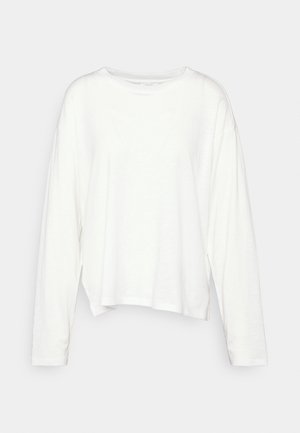 Chemise blanche à manches longues et col rond, avec une coupe décontractée et un ourlet légèrement asymétrique, présentée sur un fond clair uni.